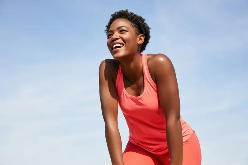 Smiling black woman in coral athletic wear bending forward outdoors under blue sky