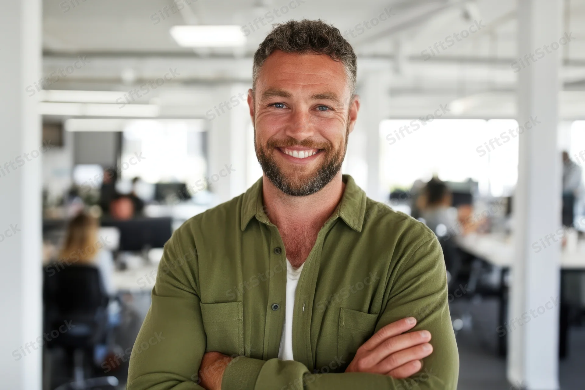 Smiling man with trimmed beard in olive green shirt standing with arms crossed in modern office workspace