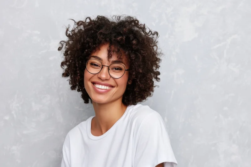 Smiling woman with tight curly hair and round glasses wearing a white t-shirt in a studio setting