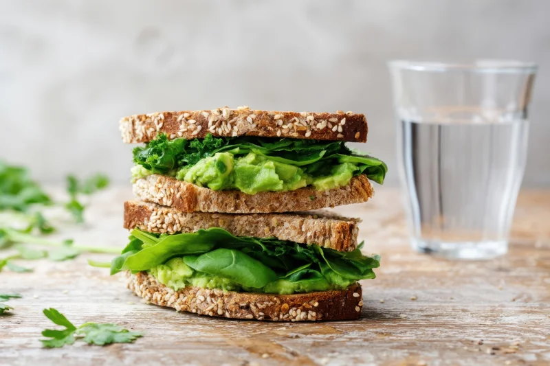 Two-tier sesame-seed crusted avocado and greens sandwich on wooden table with glass of water