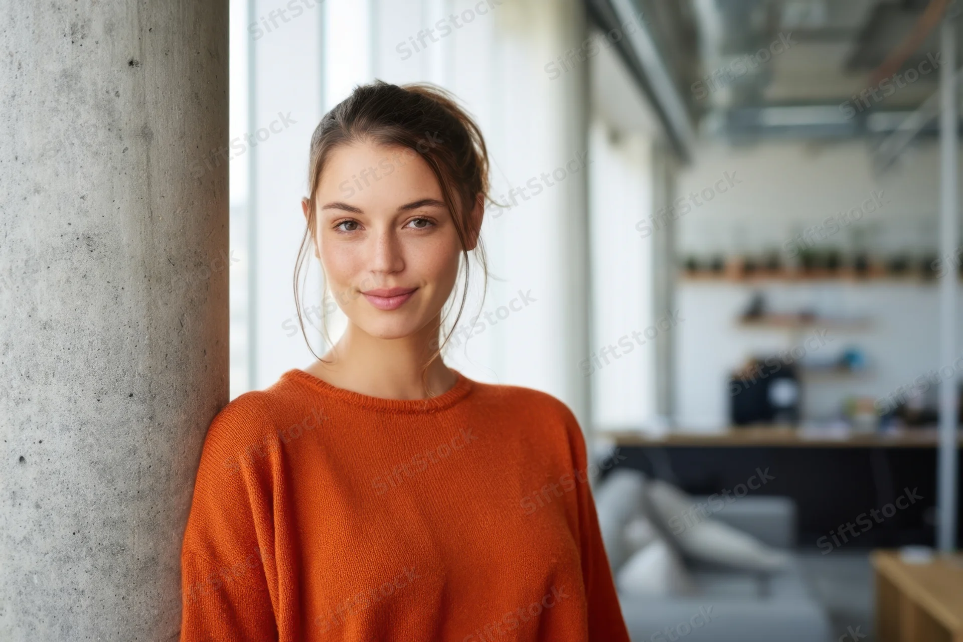 Young woman in orange sweater smiling while leaning against concrete column in modern office space