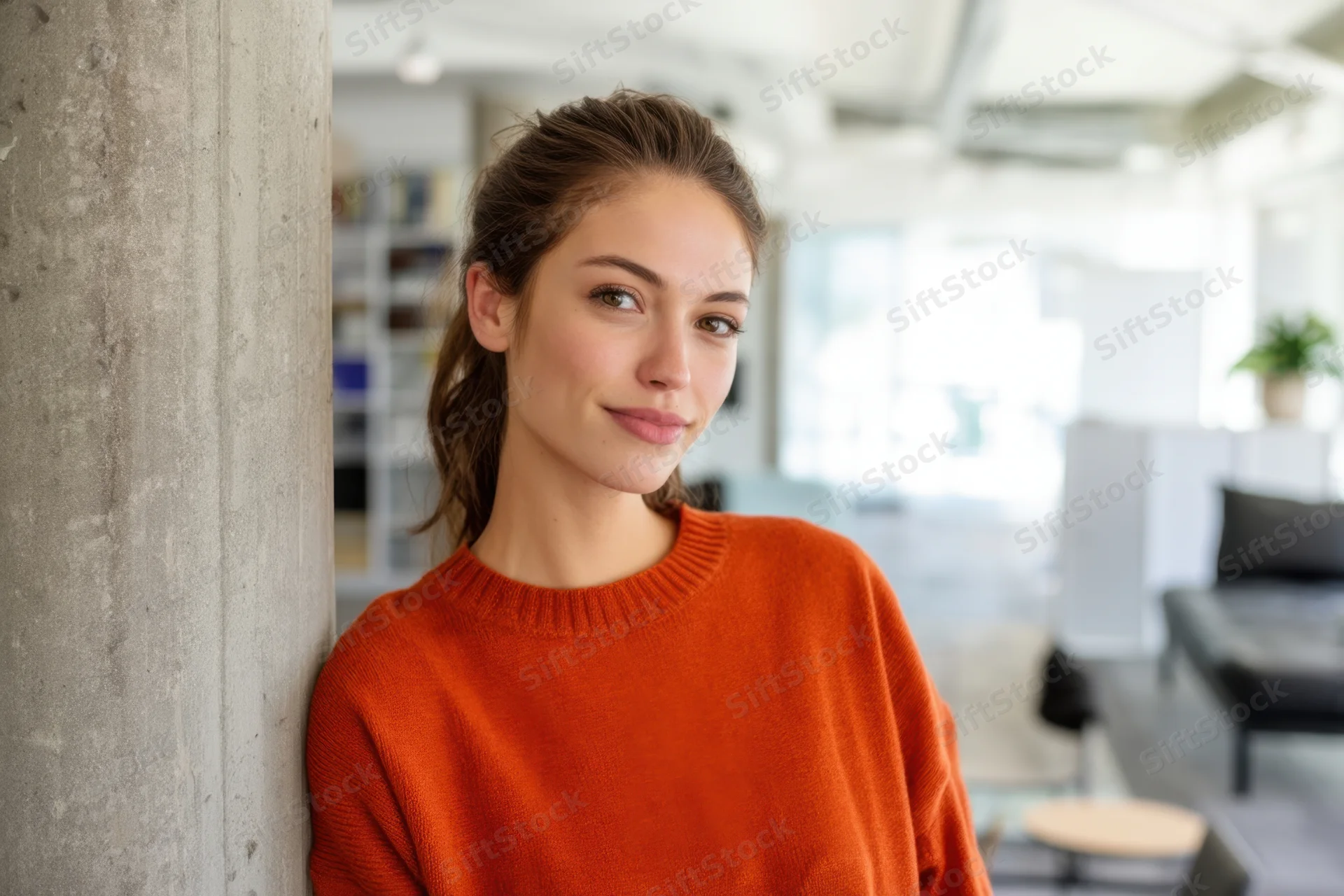 Young woman in orange sweater leaning against concrete pillar in modern office interior