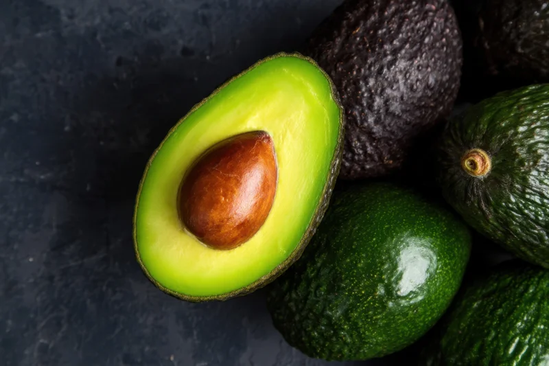 Halved avocado with pit resting among whole avocados on dark surface