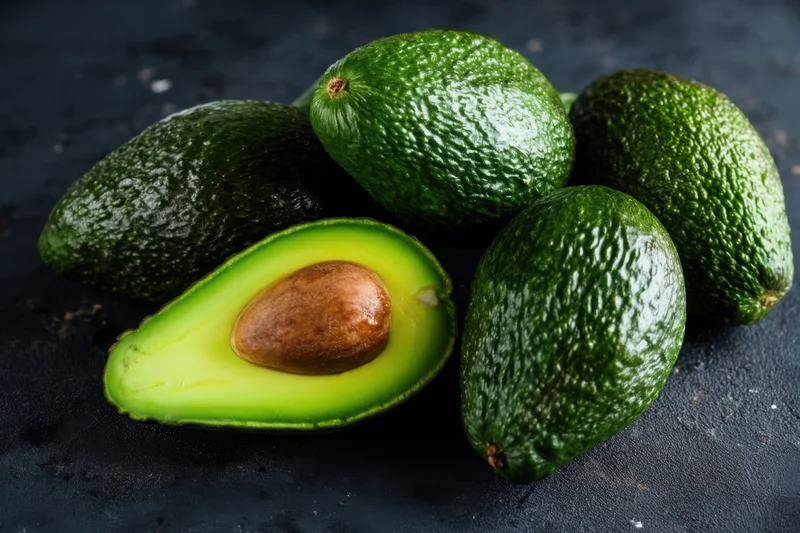 Whole avocados piled together with one half cut open to show the pit and green flesh on a dark surface