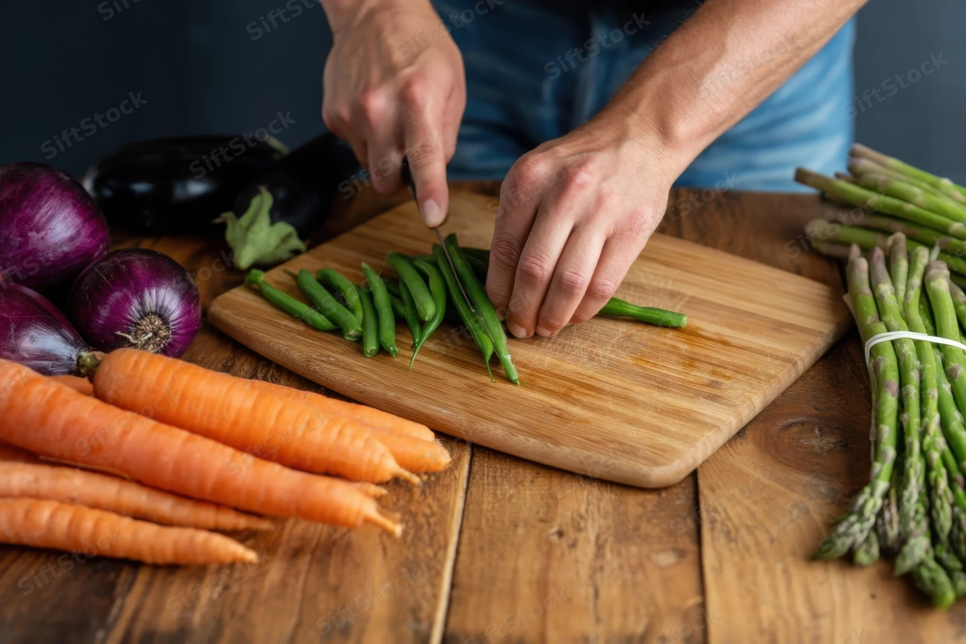 Hands slicing green beans on a wooden cutting board surrounded by fresh vegetables on a rustic table