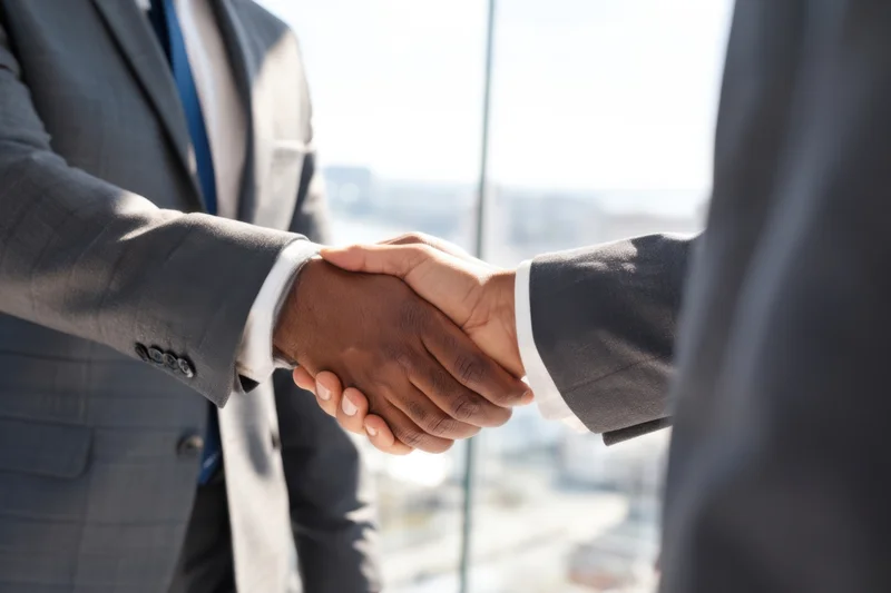 Two businessmen in suits shaking hands in a modern glass office with city view