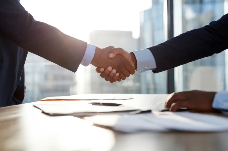 Two businessmen in suits shaking hands over a conference table in a modern office with city view