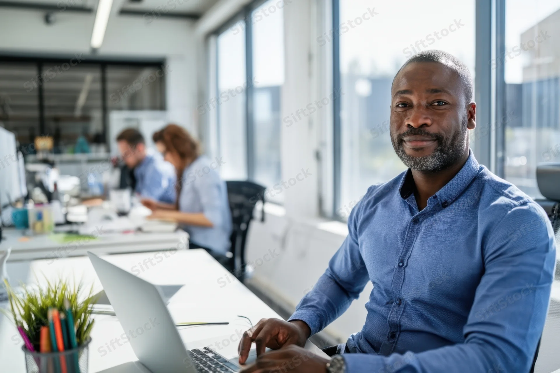 Man in blue shirt smiling at camera while working on laptop in modern open-plan office