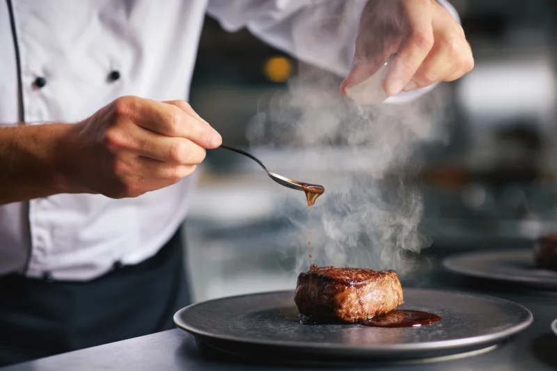 Chef plating seared steak with sauce on dark plate in restaurant kitchen