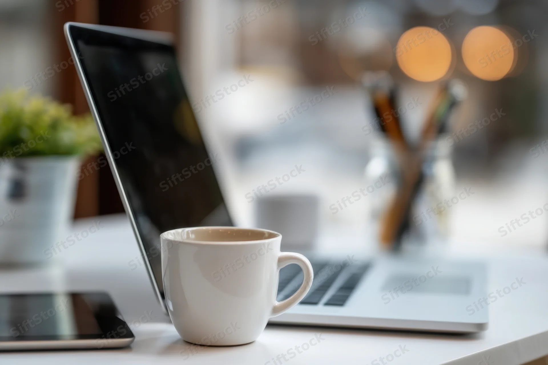 Silver laptop and white coffee mug on a clean white desk with blurred background and office supplies