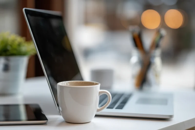Silver laptop and white coffee mug on a clean white desk with blurred background and office supplies
