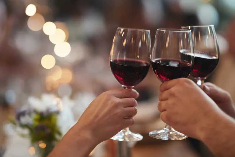 Hands holding red wine glasses toasting at a dinner table with blurred lights and flowers in the background