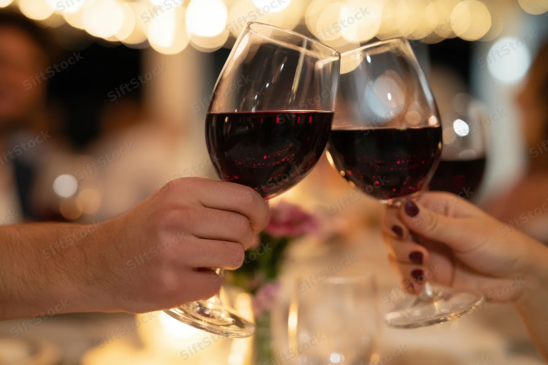 Hands clinking red wine glasses at a dinner table with candlelight and bokeh lights in the background