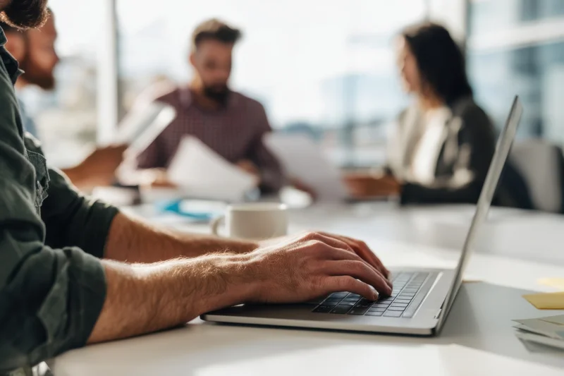 Hands typing on a laptop at a conference table with colleagues in the background