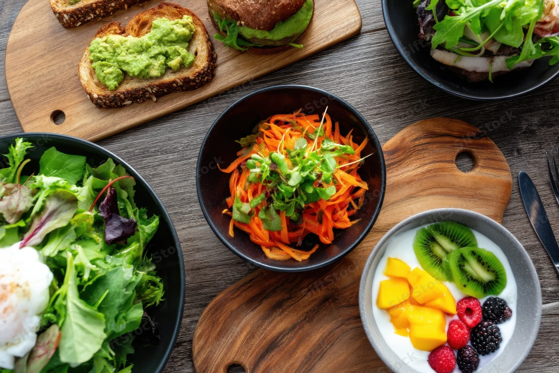 Overhead view of a brunch spread with avocado toast, salads, yogurt with fruit, and carrot salad on wooden boards and bowls on a rustic table