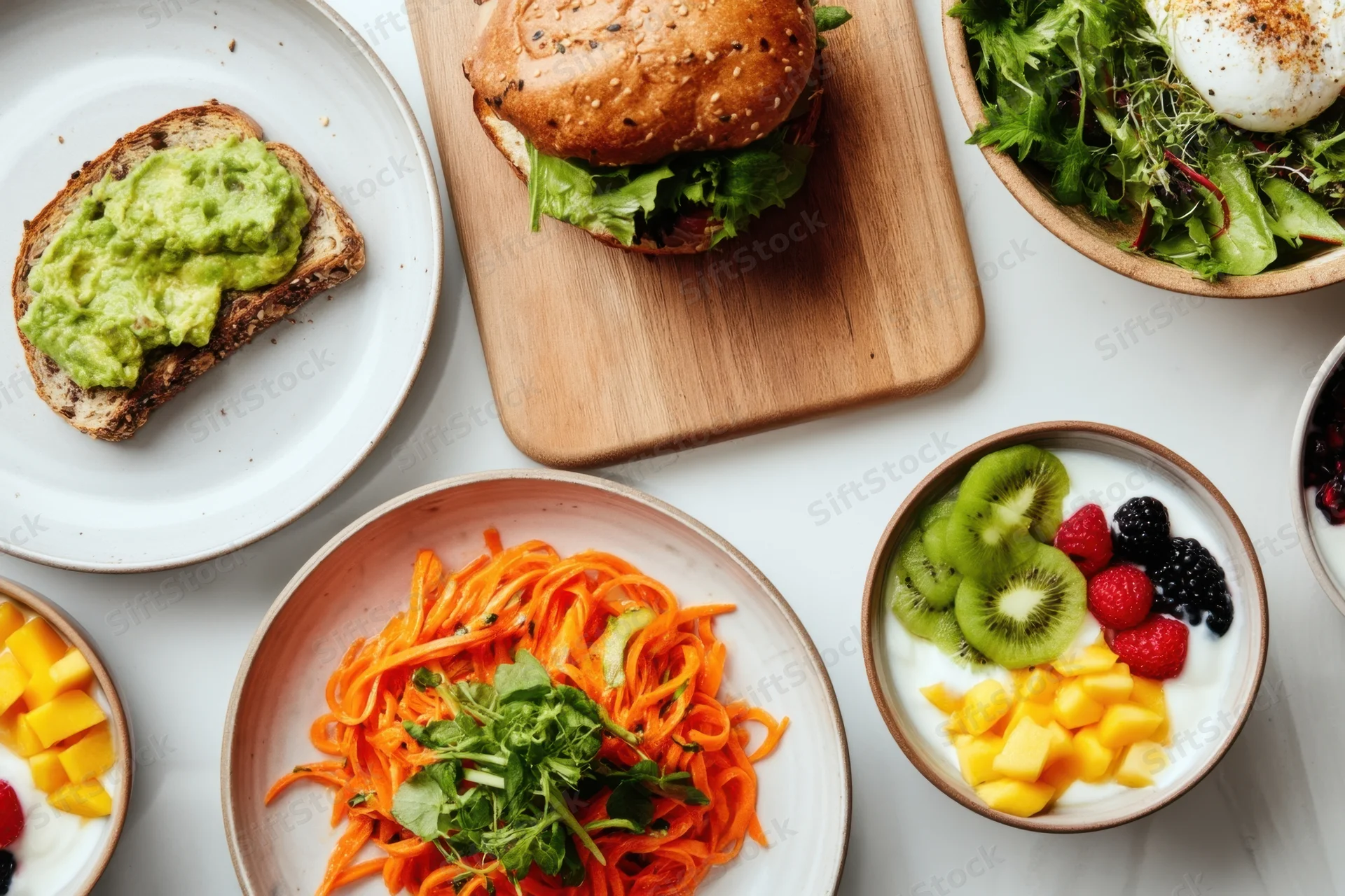 Overhead view of a brunch spread with avocado toast, sandwich, salad, yogurt with fruit, and spiralized carrots on a white table