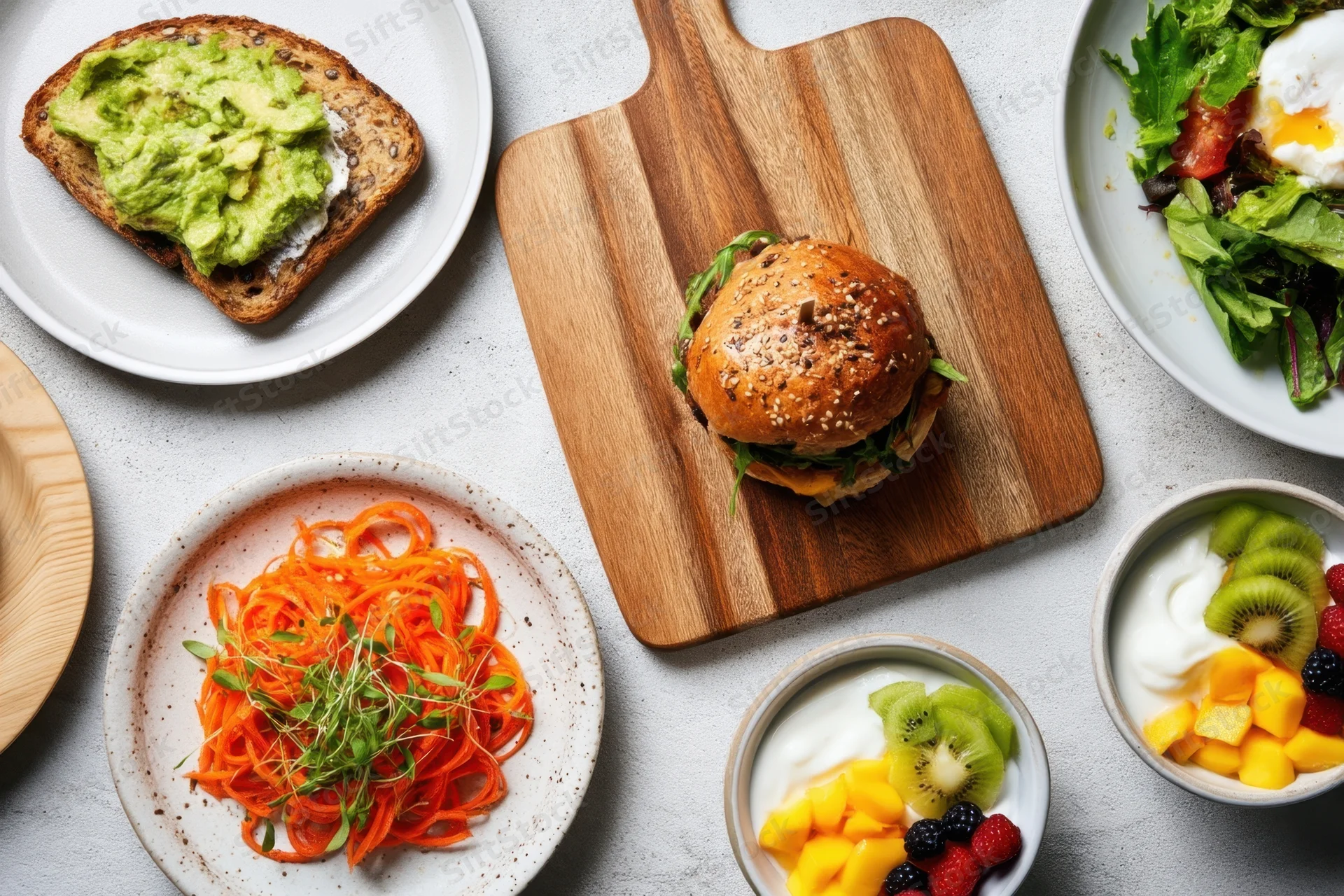 Overhead view of a brunch spread with avocado toast, burger, salad, yogurt with fruit, and spiralized carrots on a white table