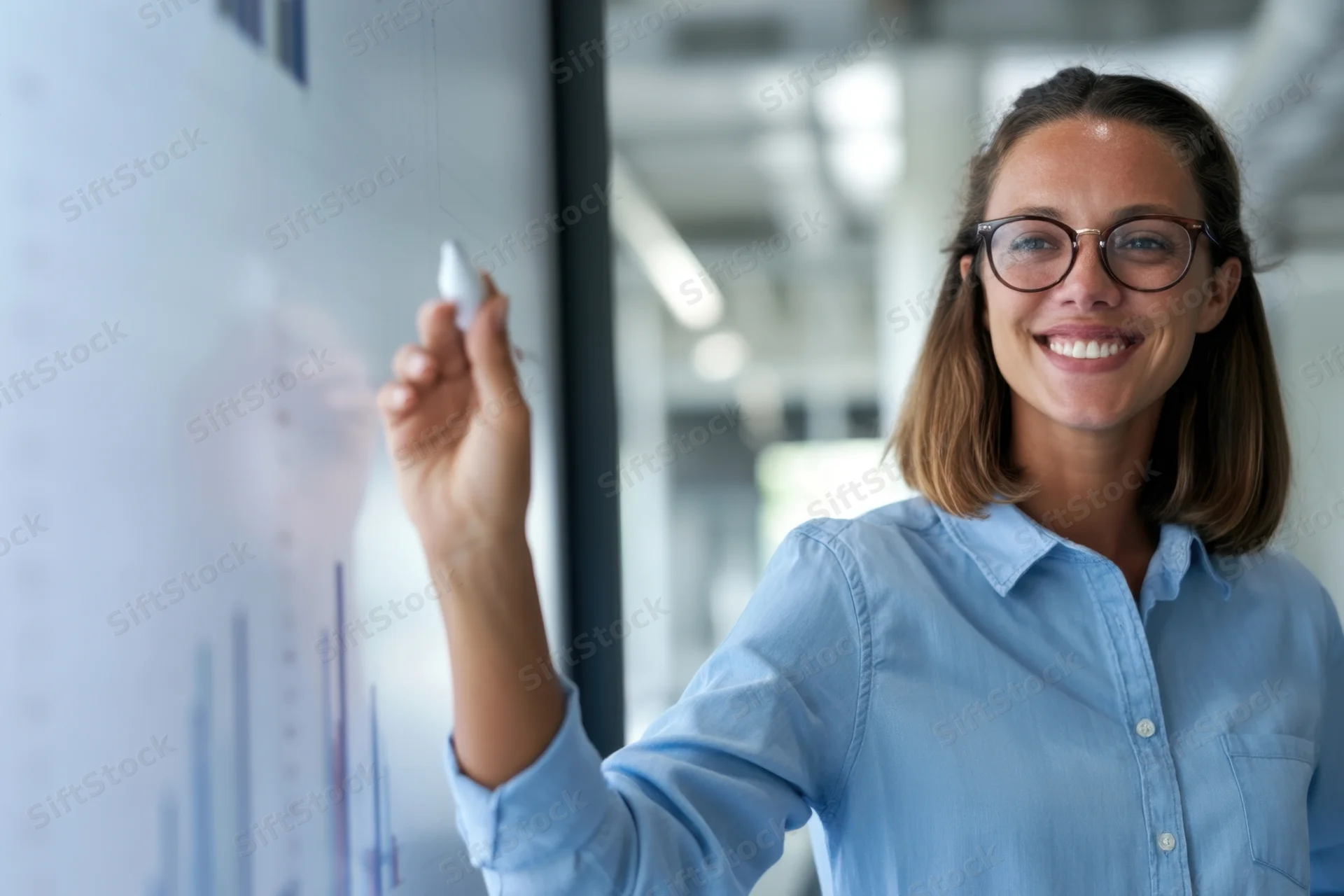 Woman in blue shirt smiling while writing on whiteboard with marker in office