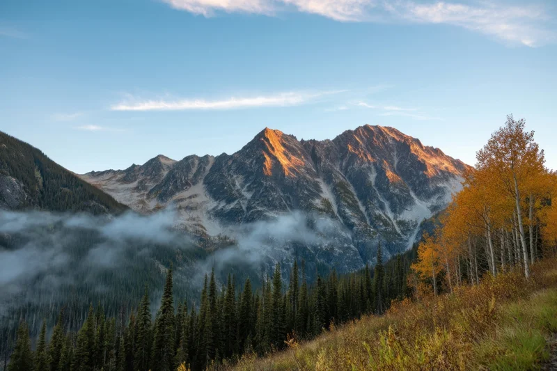Jagged granite peaks rising above a misty valley with autumn-colored aspen and pine trees under a clear blue sky