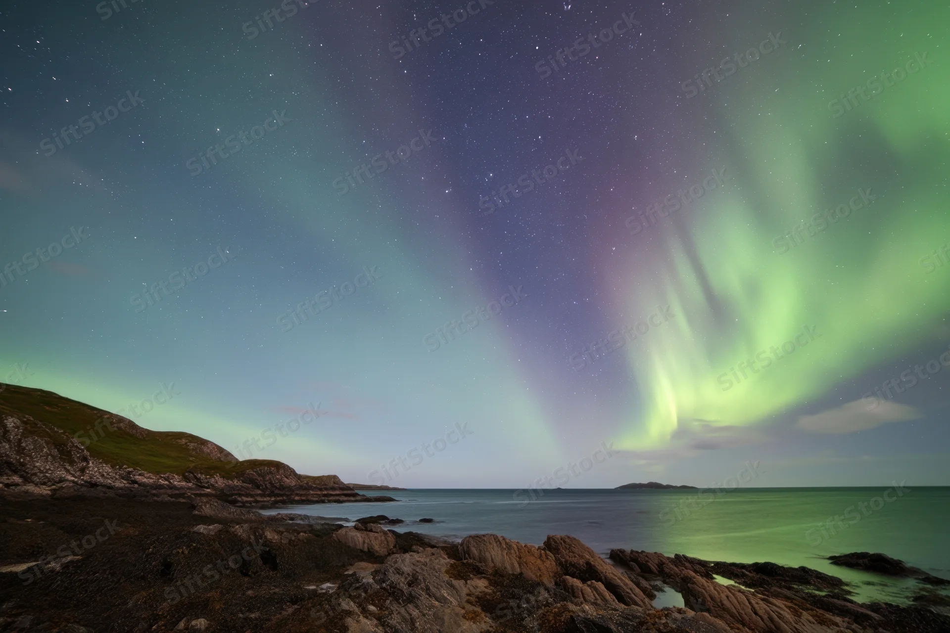 Aurora borealis glowing over rocky coastline at night with stars and ocean in view