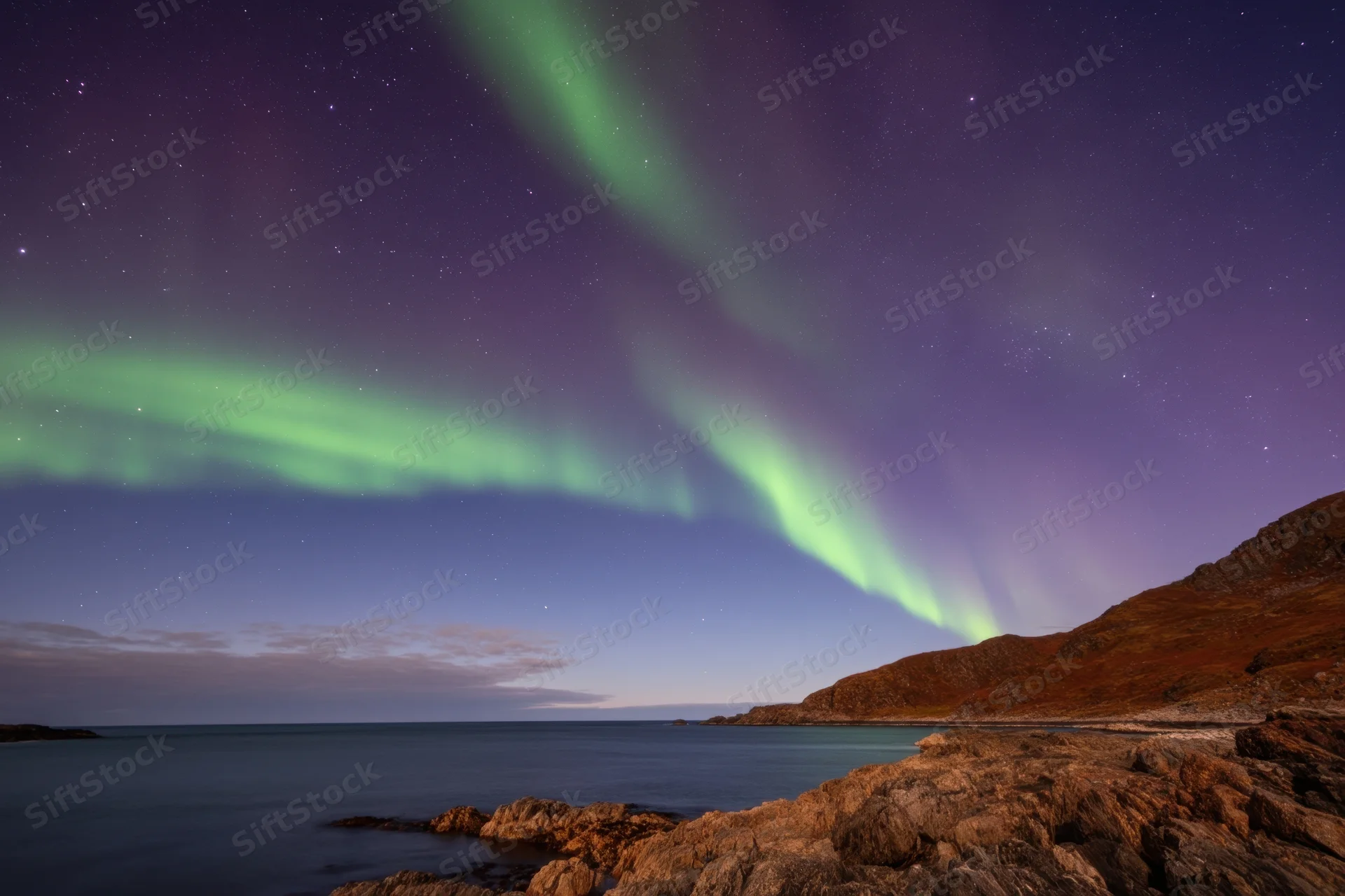 Aurora borealis glowing over rocky coastline at night with stars and ocean in view