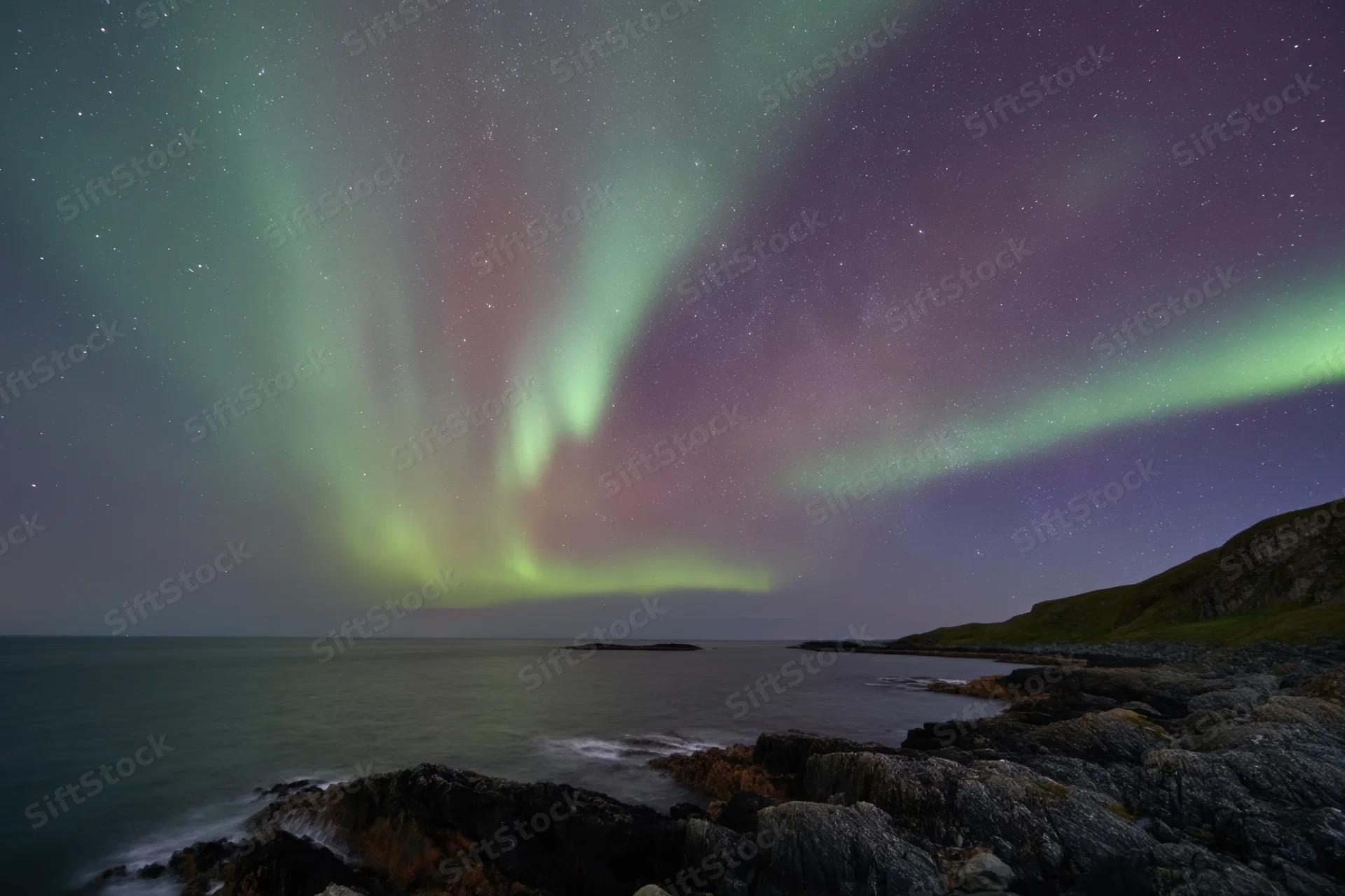 Aurora borealis glowing over rocky coastline at night with stars and ocean in view