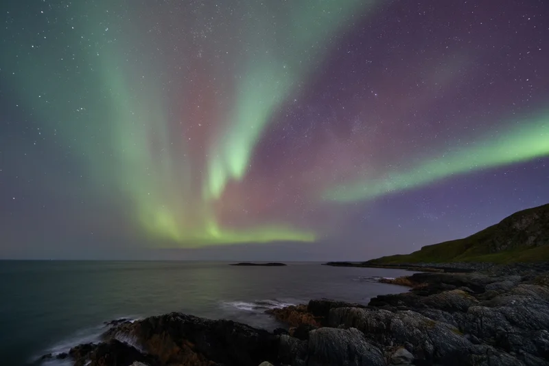 Aurora borealis glowing over rocky coastline at night with stars and ocean in view