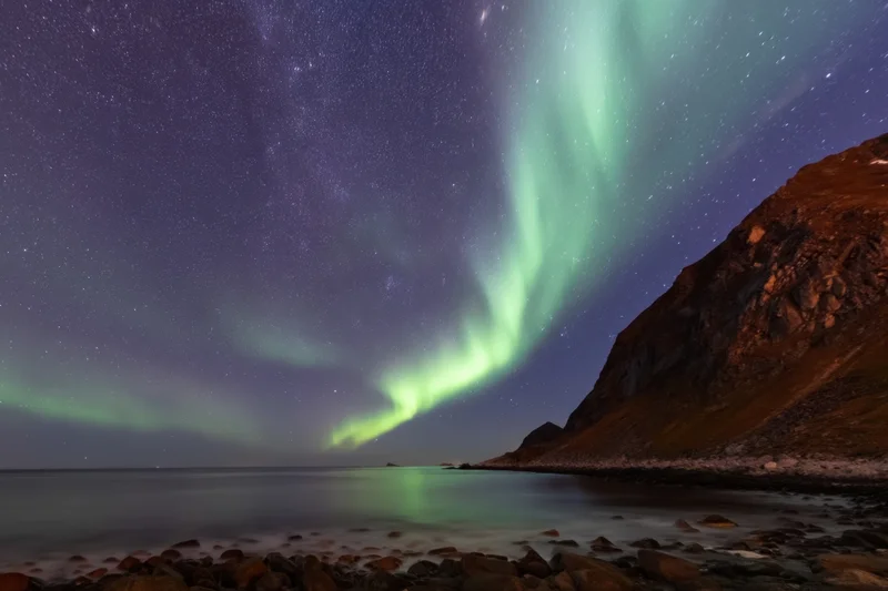 Aurora borealis glowing over rocky shore and calm sea under starry night sky