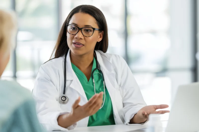 Female doctor in white coat and green scrubs explaining something to a patient at a table in a clinic office