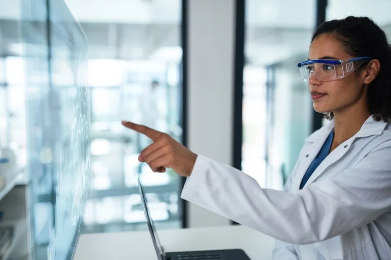 Woman in lab coat pointing at digital display in modern laboratory workspace