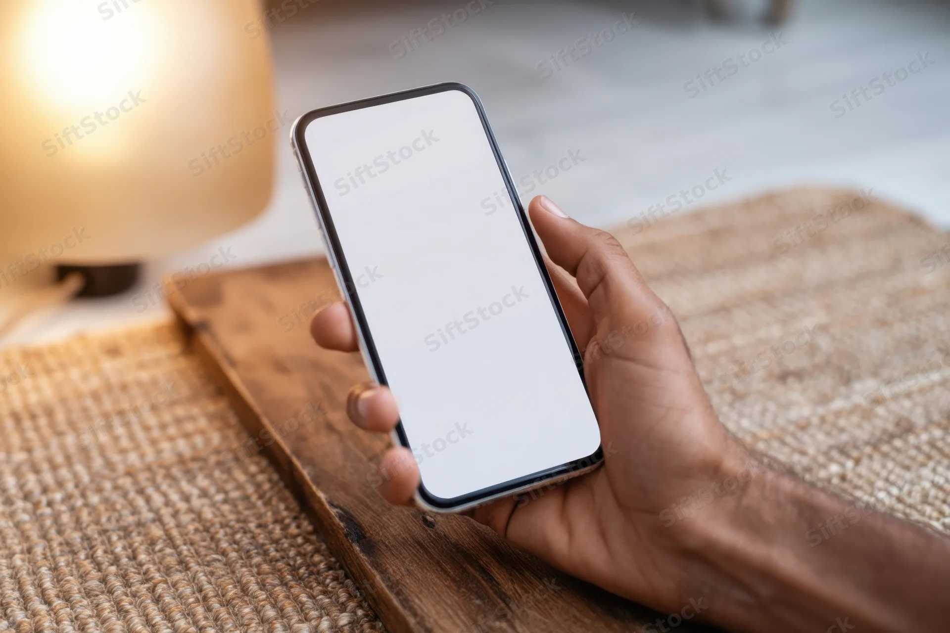 Hand holding smartphone with blank screen above wooden table in home interior