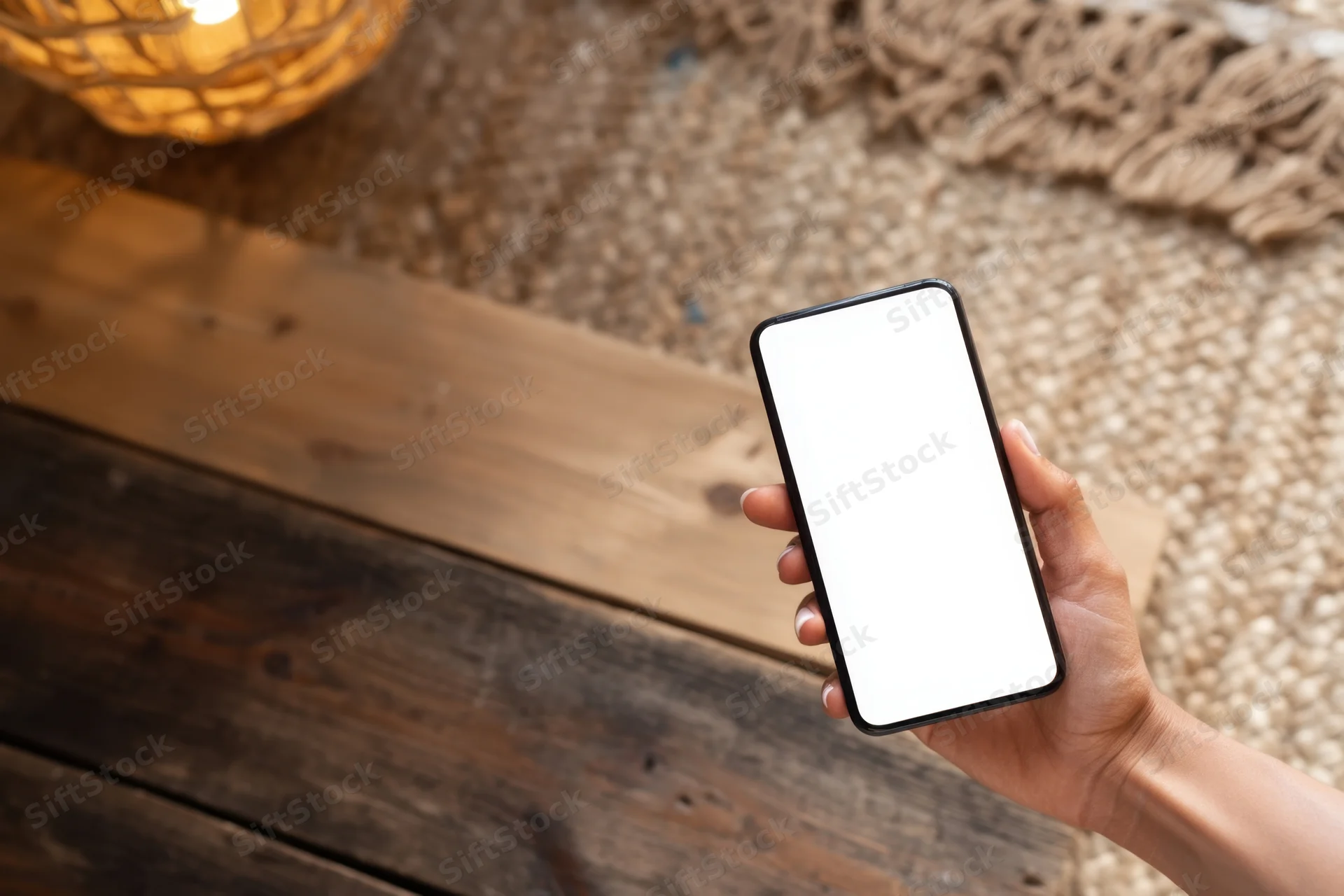 Hand holding smartphone with blank screen above wooden floor and woven rug