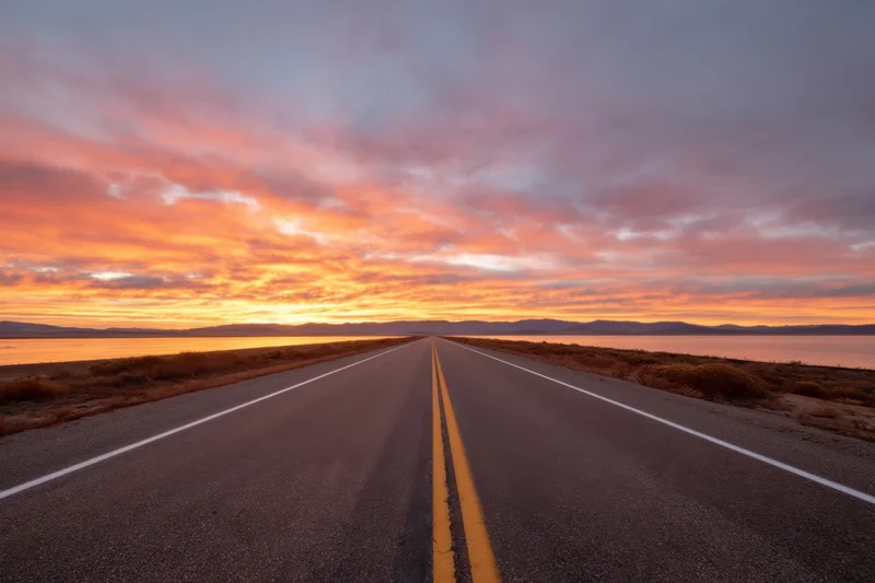 Long straight two-lane road stretching toward sunset over a calm lake with distant mountains on the horizon