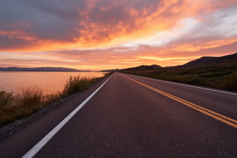 Long two-lane road stretching toward a sunset over a tranquil lake with mountains in the distance