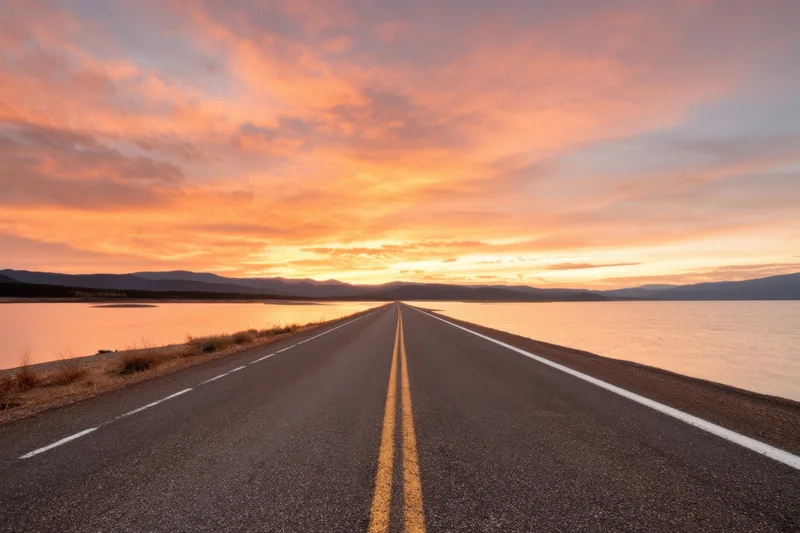 Long straight road running along a calm lake under a vibrant sunset sky