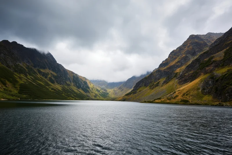 Calm dark lake in a moody alpine valley flanked by rugged mountains under overcast sky