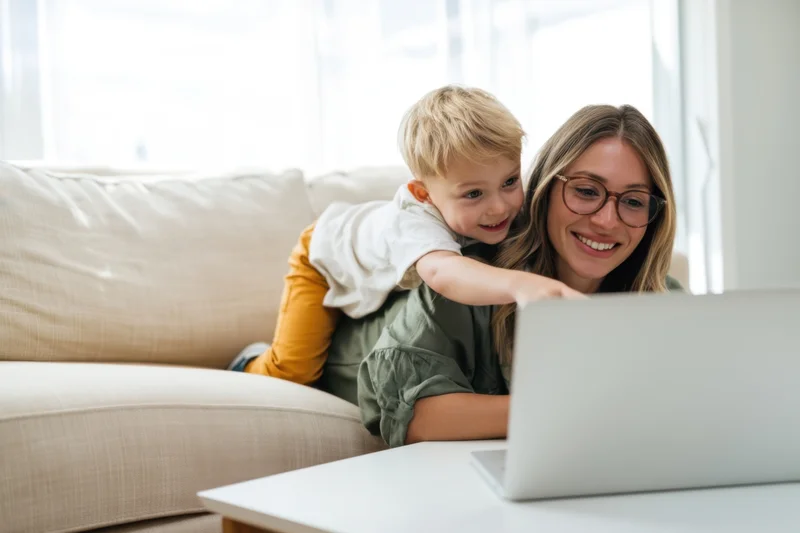 Mother smiling with toddler on lap while using laptop on sofa at home