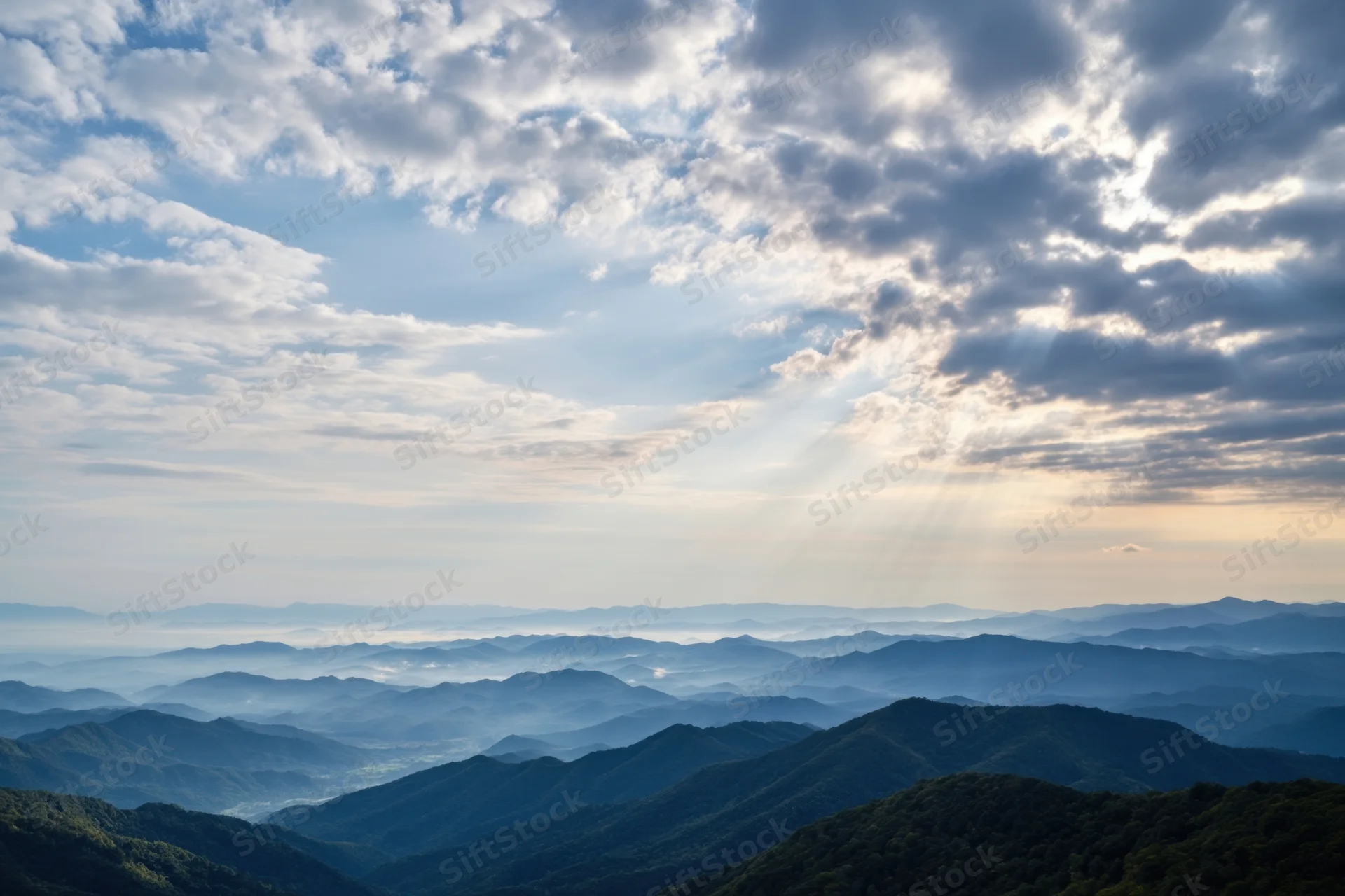 Panoramic view of layered blue mountain ridges under a cloudy sky with sunbeams breaking through