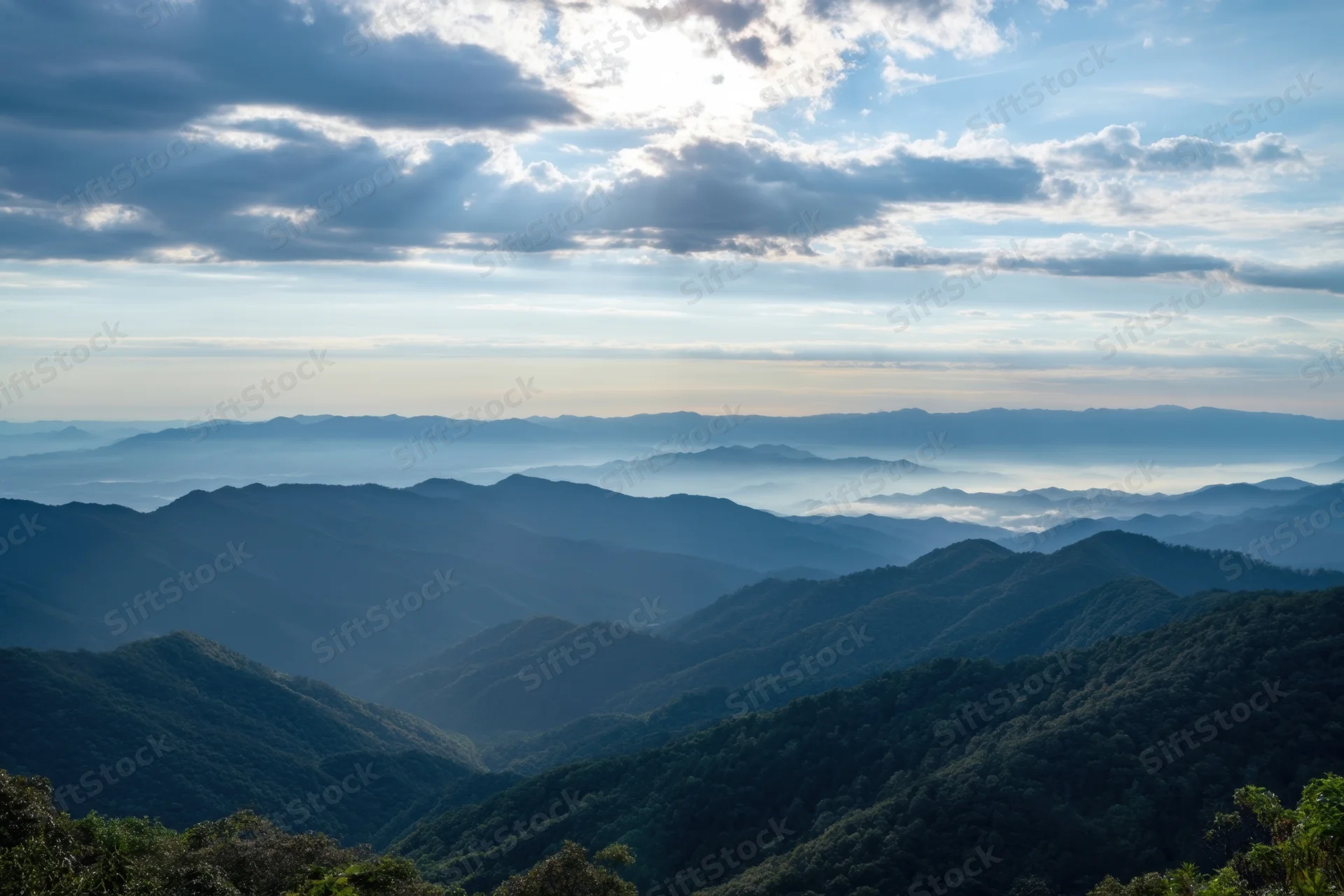 Layered blue mountain ridges covered in forest under a cloudy sky with sunlight breaking through