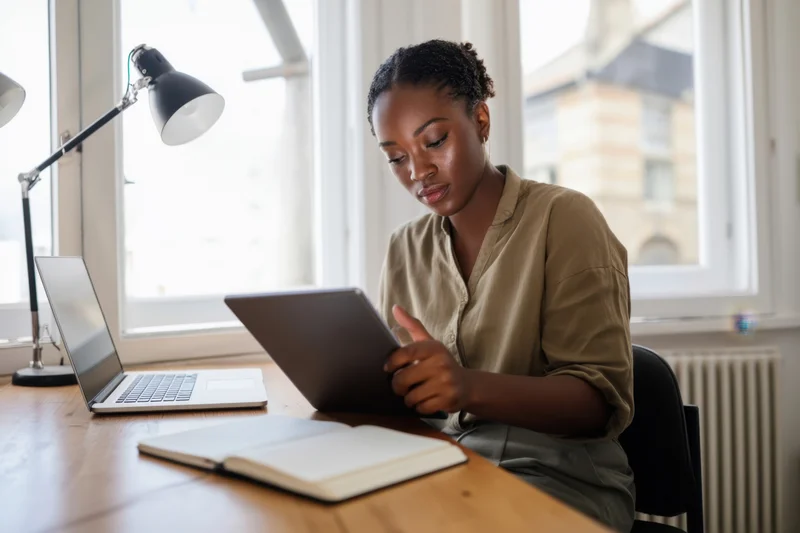 Woman tapping tablet at wooden desk with laptop and notebook nearby