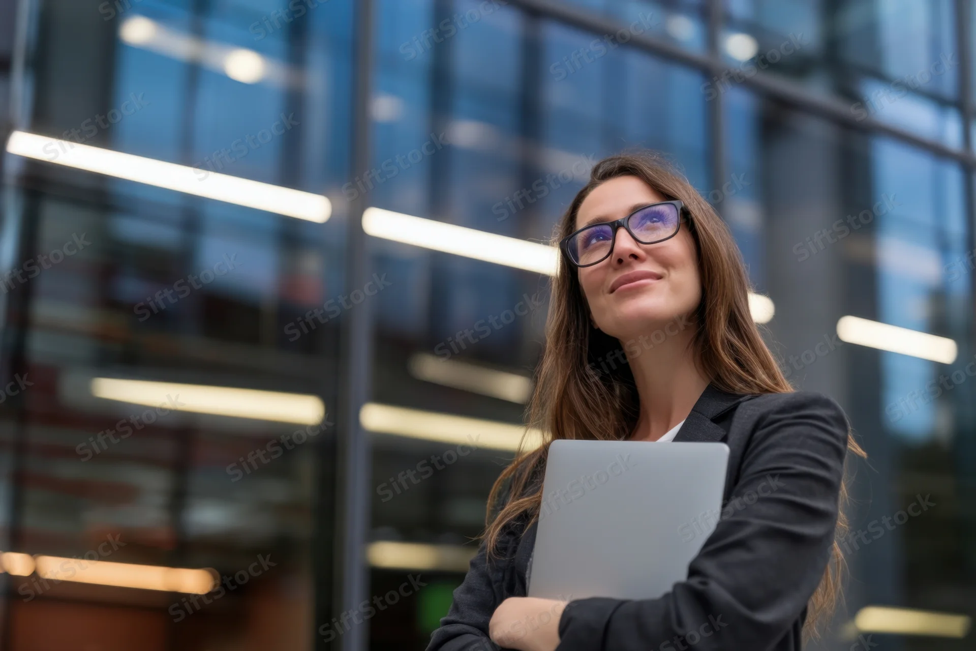 Woman in black suit holding laptop outside modern glass office building
