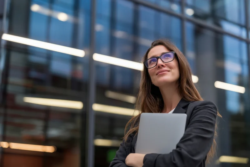 Woman in black suit holding laptop outside modern glass office building