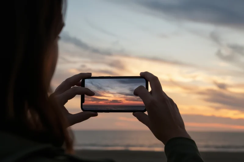 Woman holding smartphone to capture sunset over ocean from beach