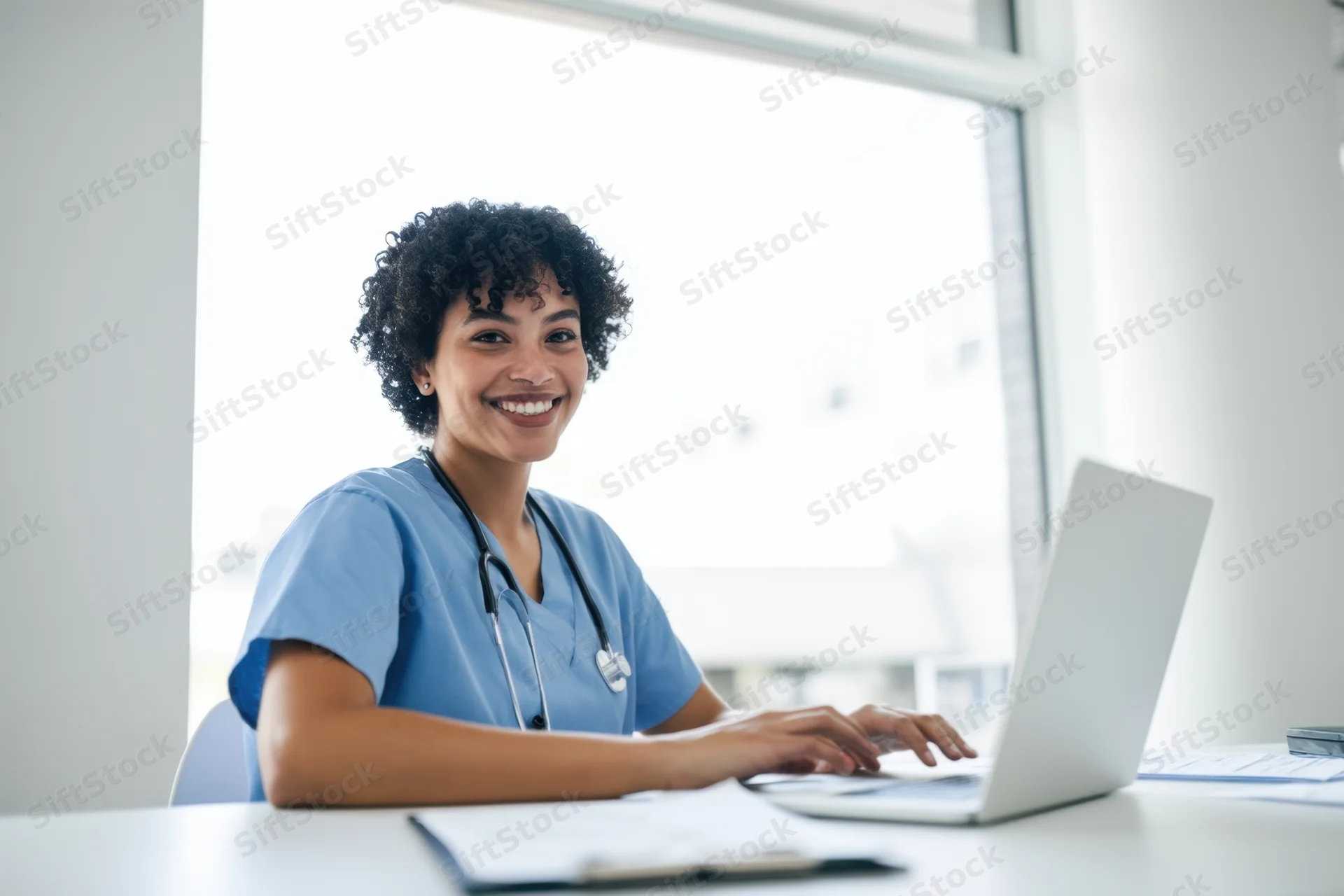 Smiling nurse in blue scrubs using laptop at desk in bright office
