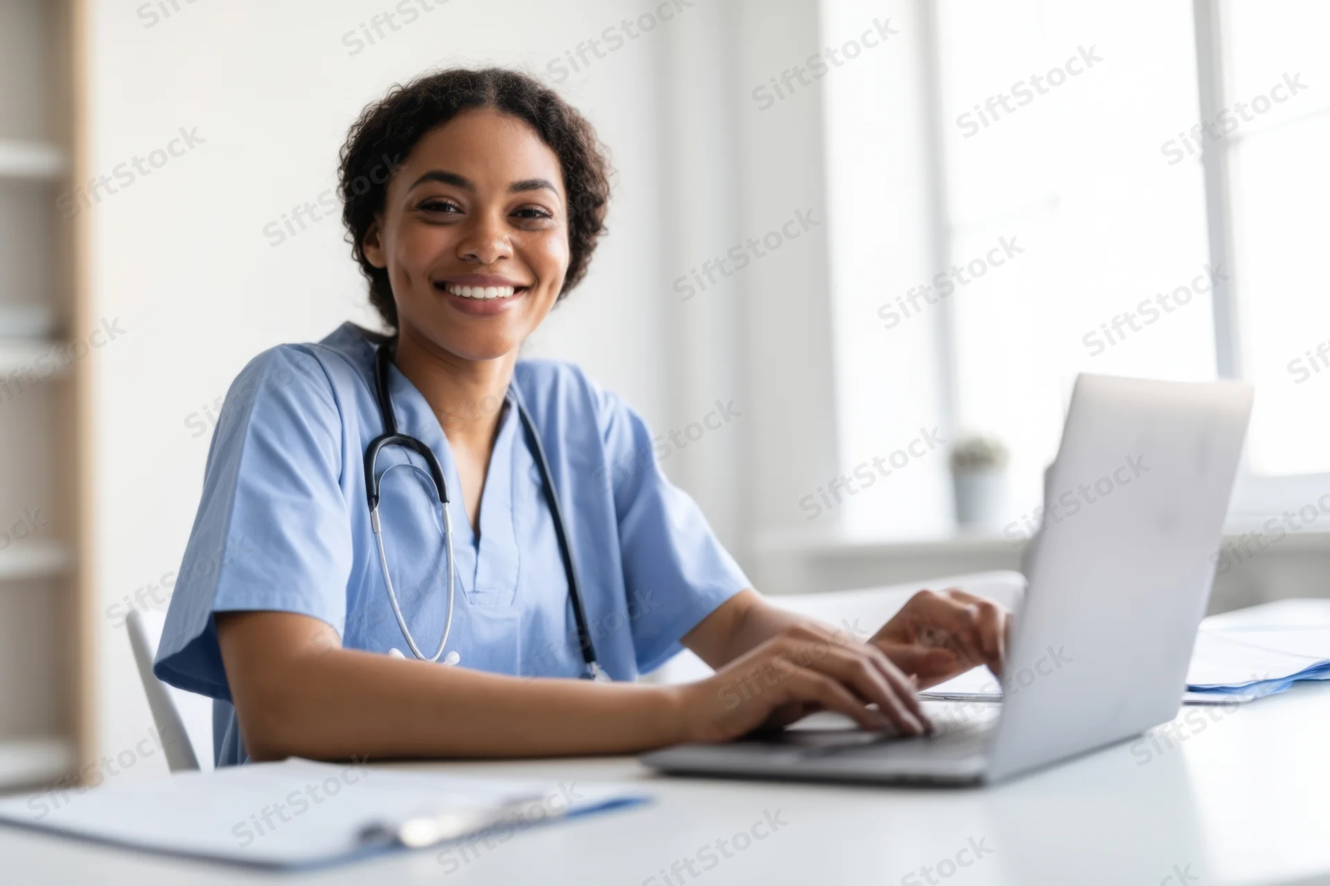 Smiling nurse in blue scrubs typing on laptop at desk in office