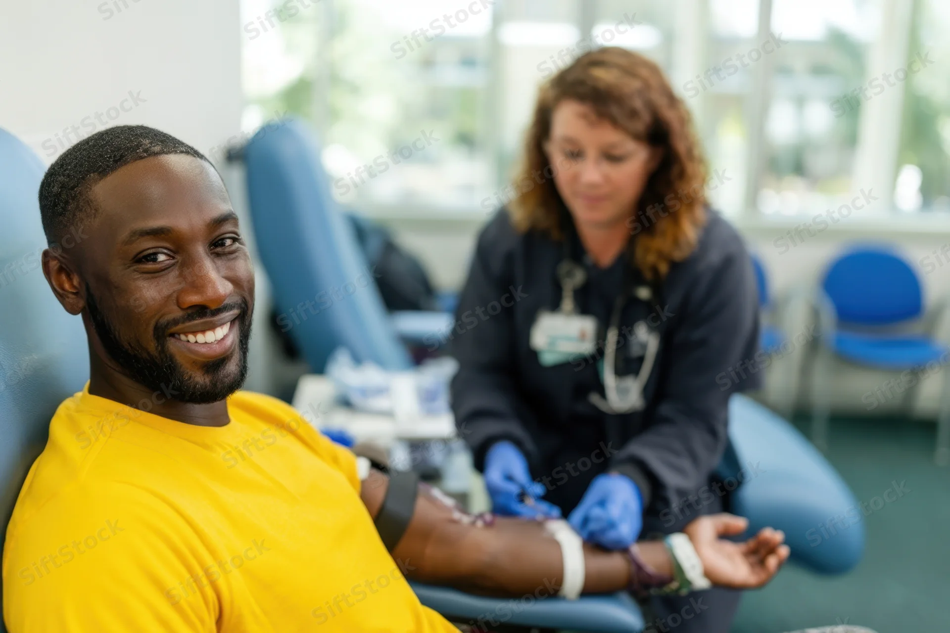 Smiling man in yellow shirt seated in donor chair during blood donation with nurse in background
