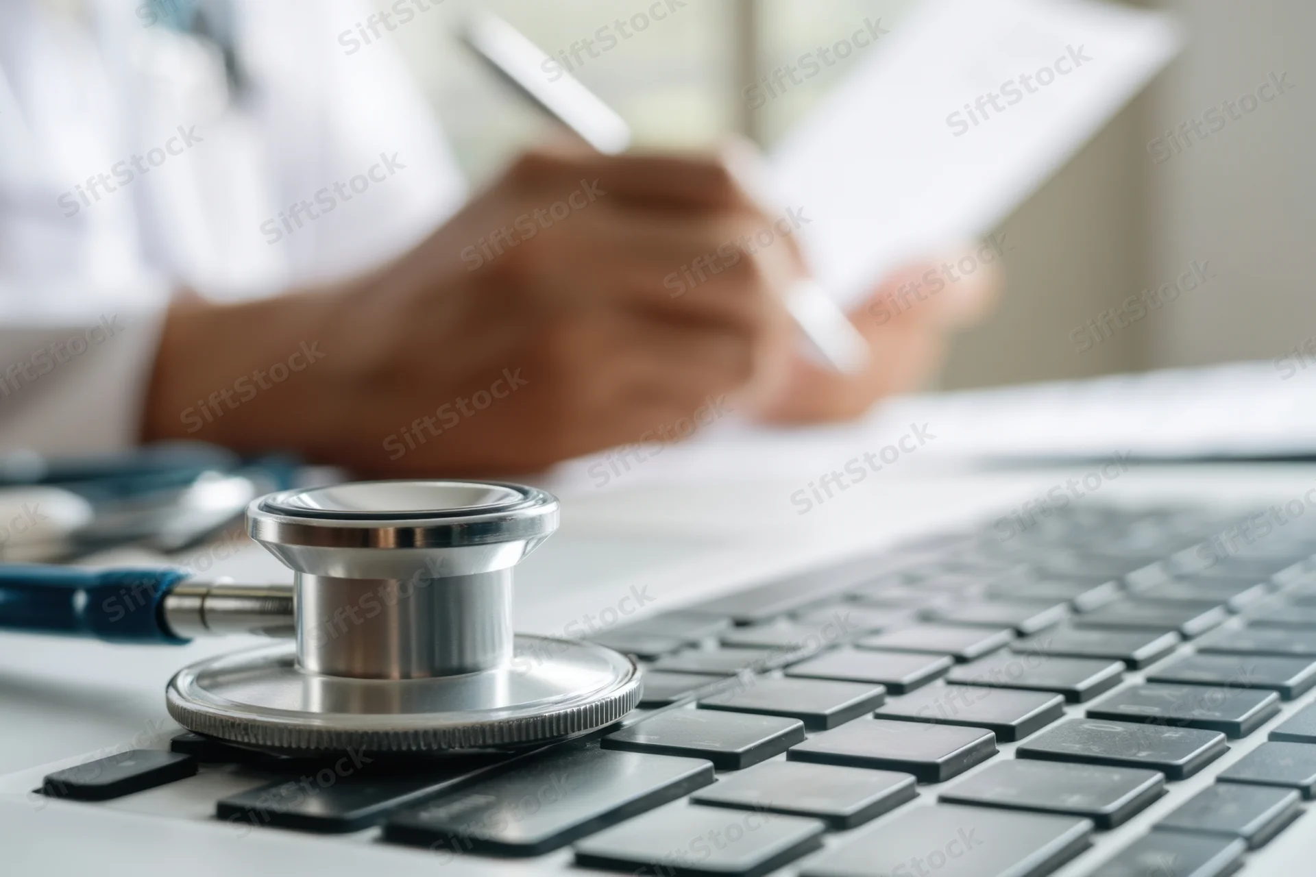 Stethoscope resting on laptop keyboard with clinician writing notes in background