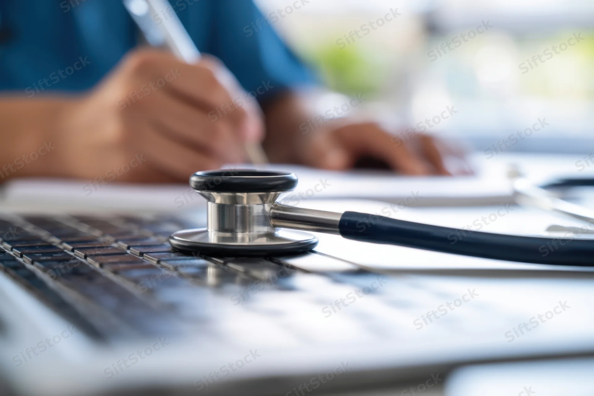 Stethoscope resting on laptop keyboard while clinician writes notes at desk