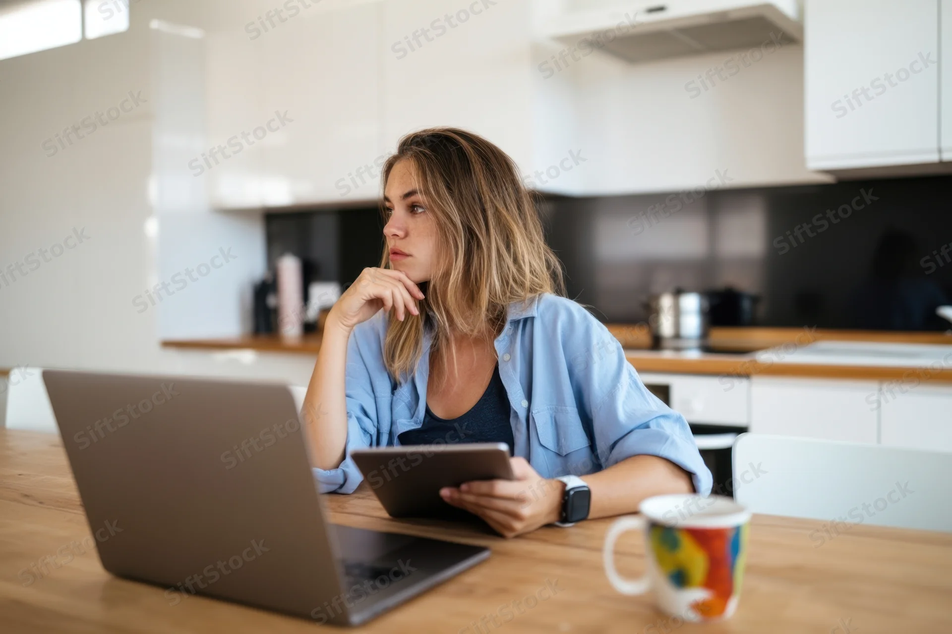 Woman sitting at wooden table in modern kitchen using laptop and tablet, looking thoughtful