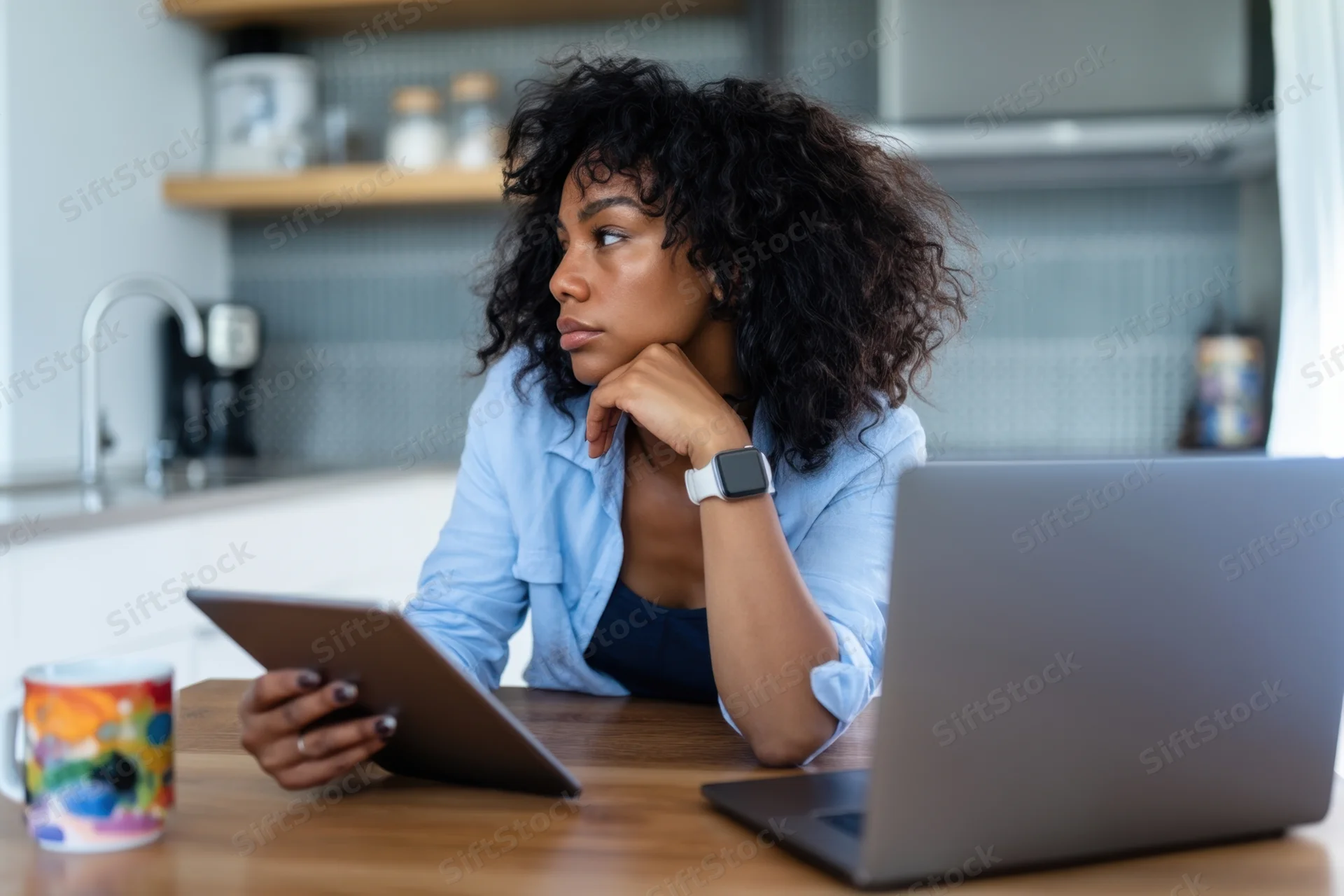 Woman sitting at wooden table in modern kitchen using tablet and laptop, thinking with hand on chin